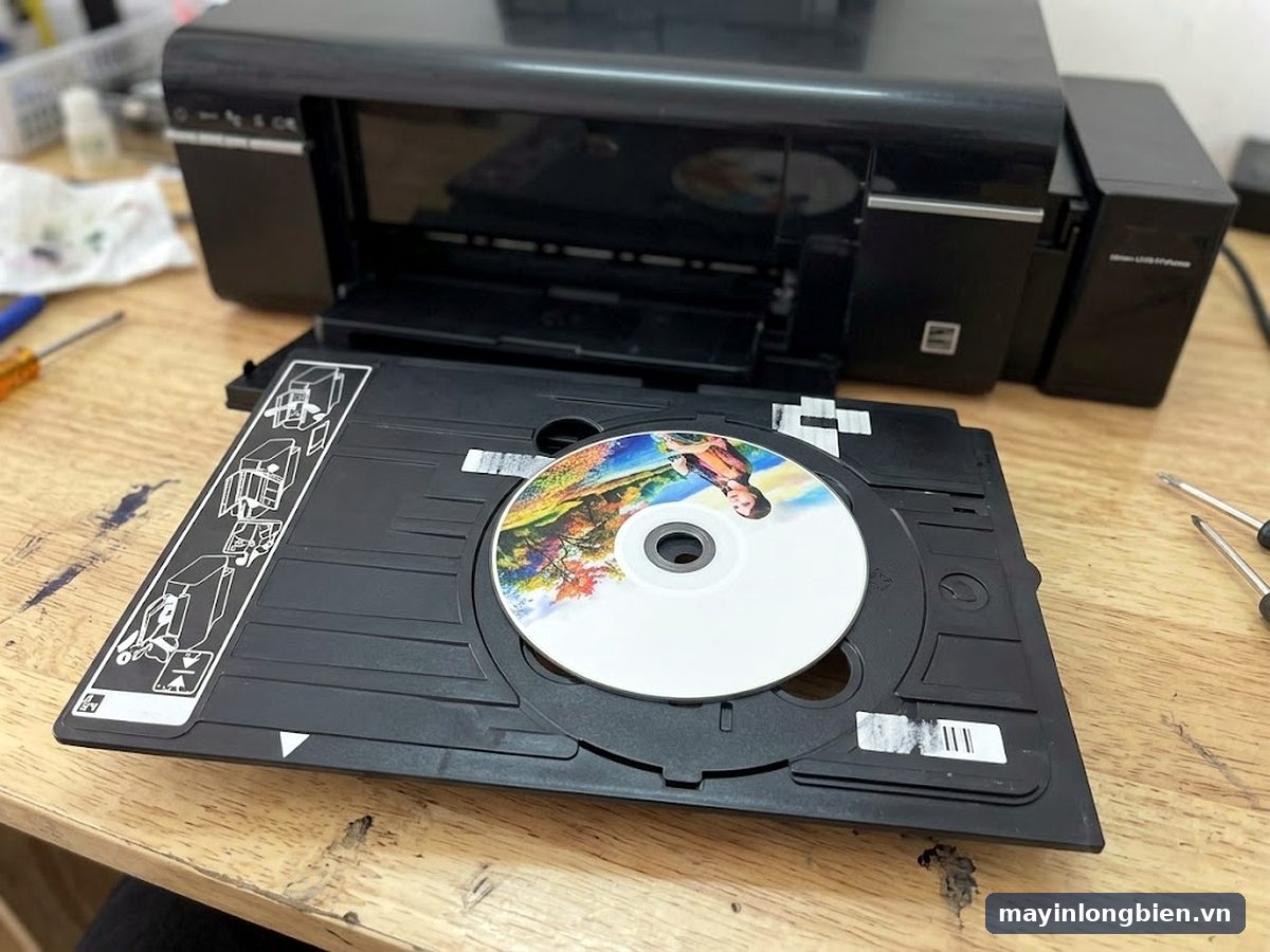 Khay in đĩa CD đặt trên bàn cạnh máy in phun A4 màu đen, đĩa in ảnh màu